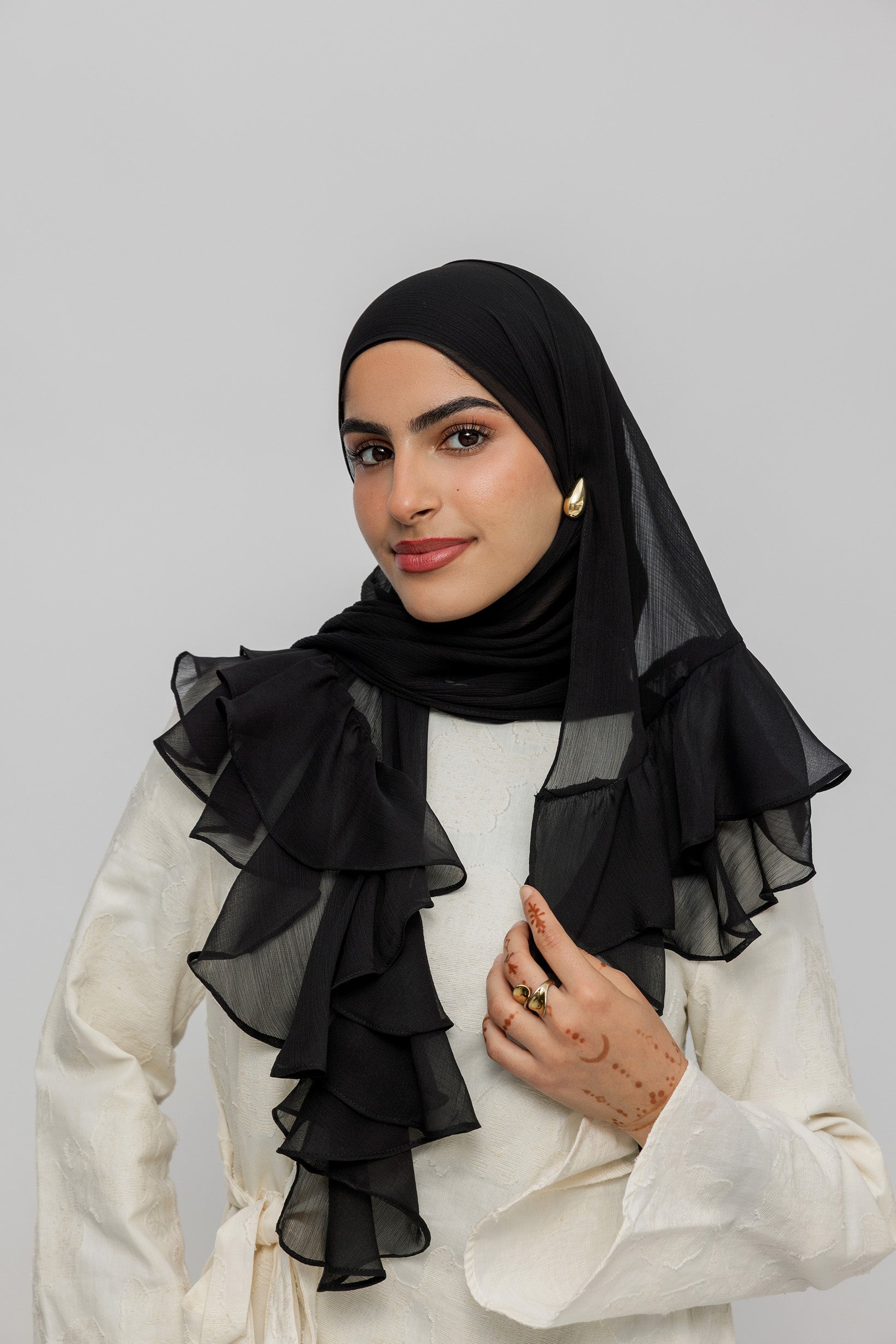 A woman wearing a black sheer ruffled hijab and a cream-colored top, posing against a solid light gray background.
