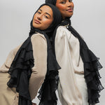 Two women modeling black ruffled hijabs and matching layered sheer shawls over neutral-toned tops, posed against a plain light gray studio background.