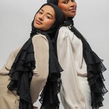 Two women modeling black ruffled hijabs and matching layered sheer shawls over neutral-toned tops, posed against a plain light gray studio background.