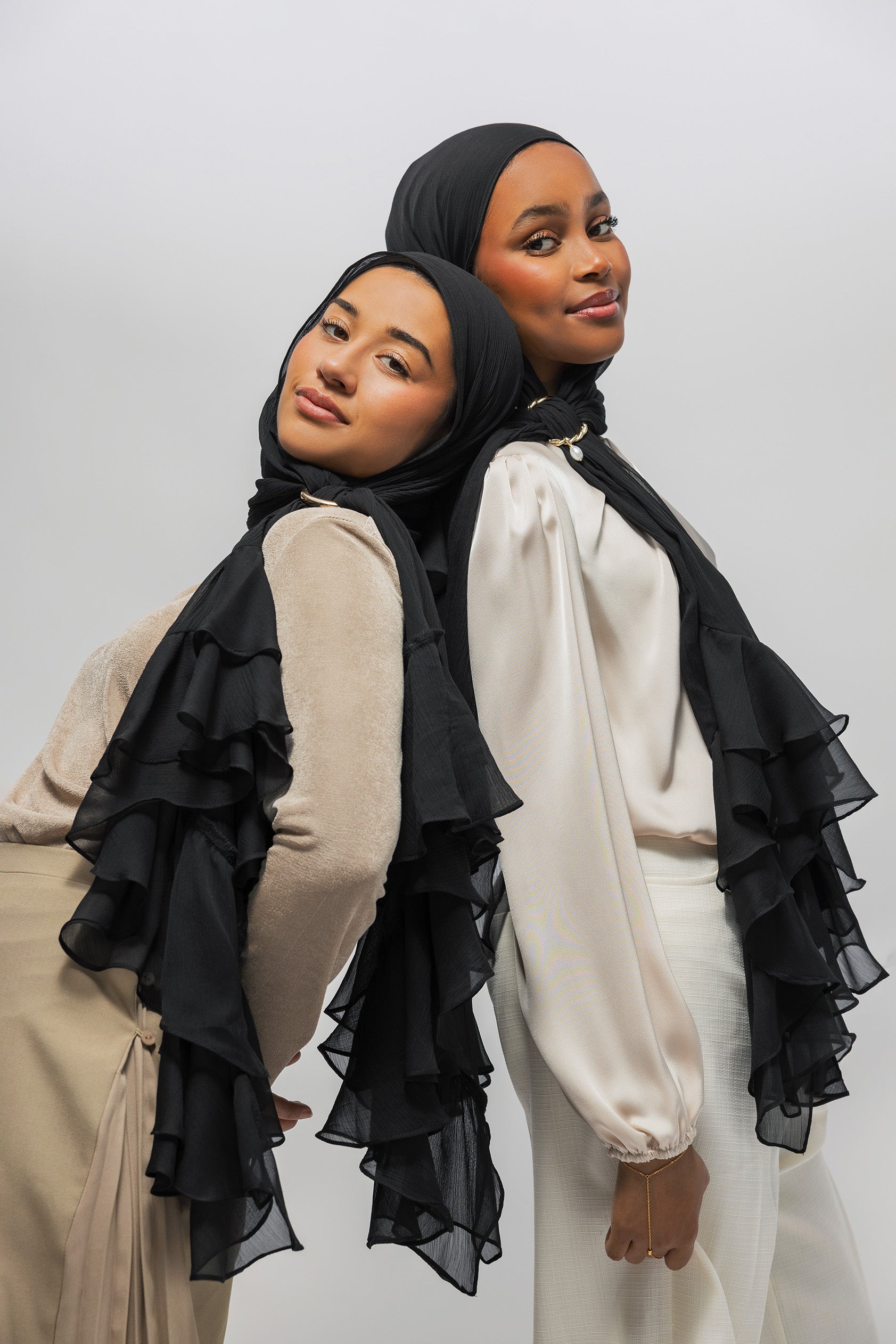 Two women modeling black ruffled hijabs and matching layered sheer shawls over neutral-toned tops, posed against a plain light gray studio background.