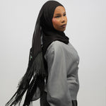 A woman wearing a black hijab with fringe trim and a light gray long-sleeve top, shown from the side against a plain light gray studio background.