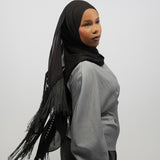 A woman wearing a black hijab with fringe trim and a light gray long-sleeve top, shown from the side against a plain light gray studio background.