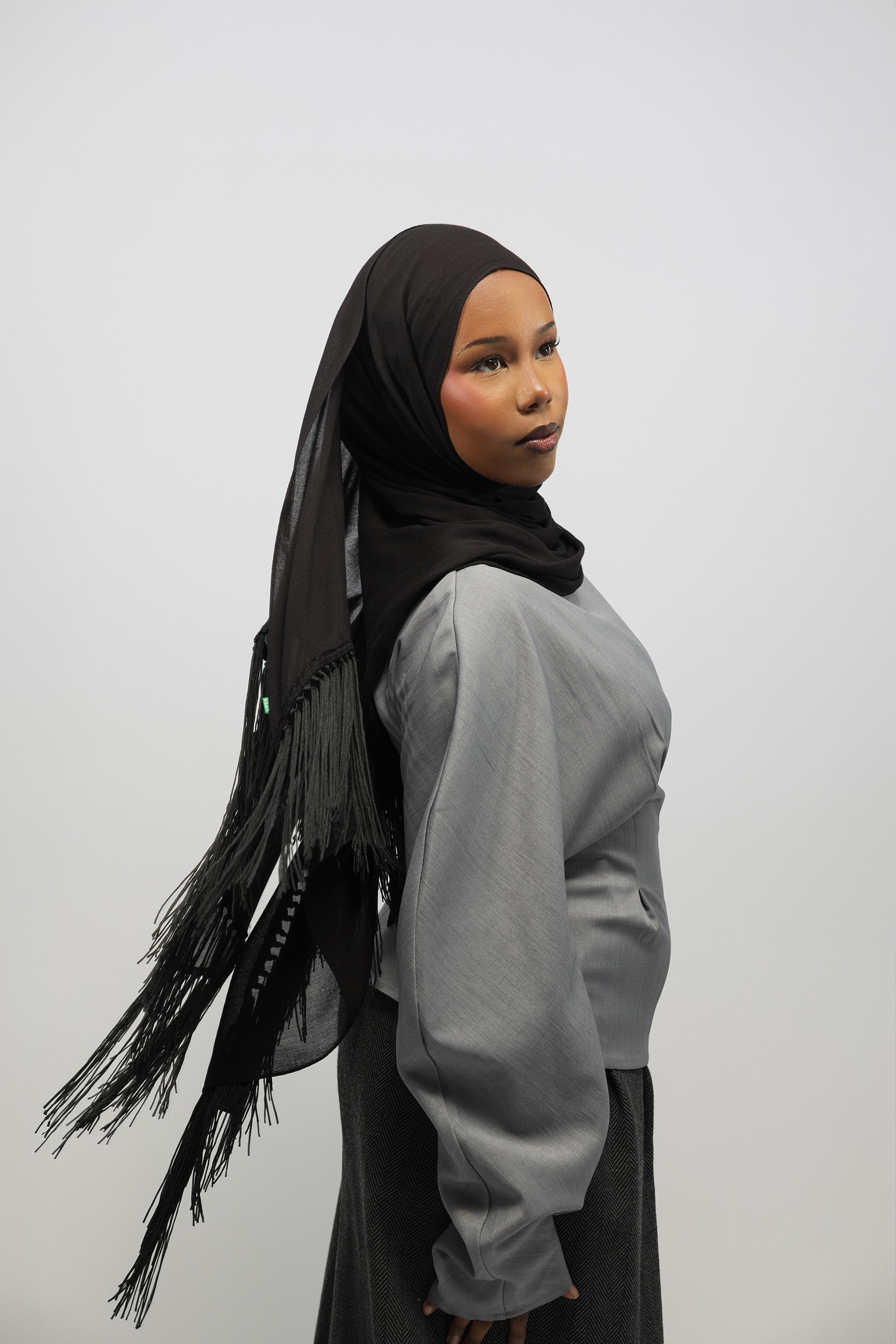 A woman wearing a black hijab with fringe trim and a light gray long-sleeve top, shown from the side against a plain light gray studio background.