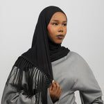 A woman wearing a black hijab with long fringe detailing, paired with a light gray long-sleeve top, photographed against a neutral light gray background.