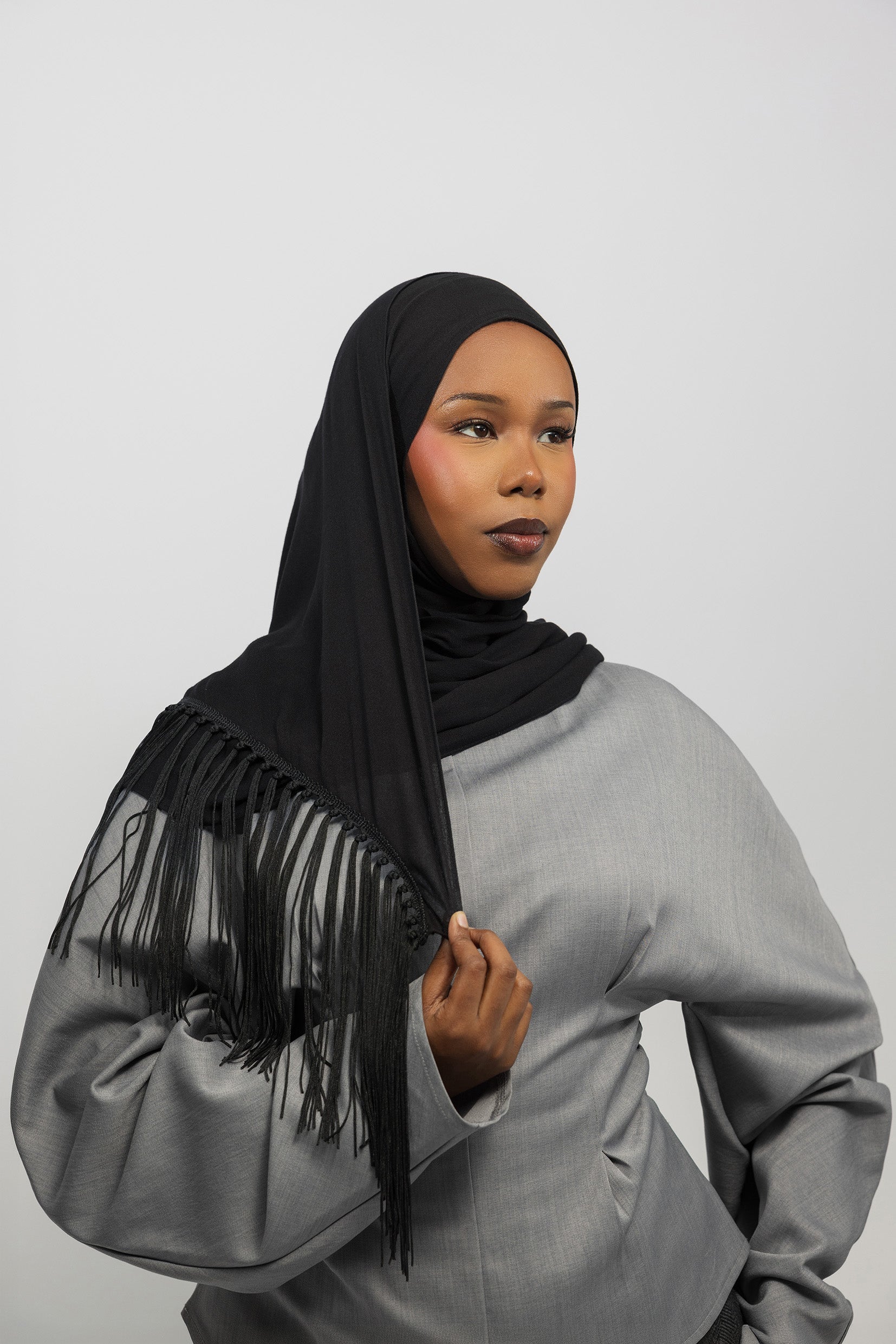 A woman wearing a black hijab with long fringe detailing, paired with a light gray long-sleeve top, photographed against a neutral light gray background.