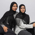 Two women wearing black fringe shawls over light-colored tops, seated against a plain light gray studio background.