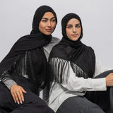 Two women wearing black fringe shawls over light-colored tops, seated against a plain light gray studio background.