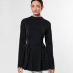 A woman modeling a black long-sleeve mock-neck top with a drawstring waist detail, paired with white pants, against a plain light gray studio background.
