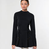 A woman modeling a black long-sleeve mock-neck top with a drawstring waist detail, paired with white pants, against a plain light gray studio background.