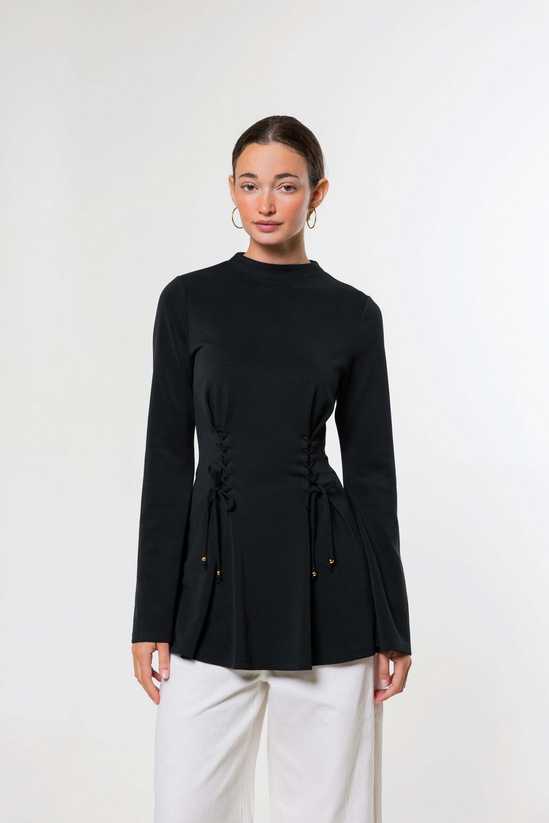 A woman modeling a black long-sleeve mock-neck top with a drawstring waist detail, paired with white pants, against a plain light gray studio background.