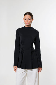 A woman modeling a black long-sleeve mock-neck top with a drawstring waist detail, paired with white pants, against a plain light gray studio background.