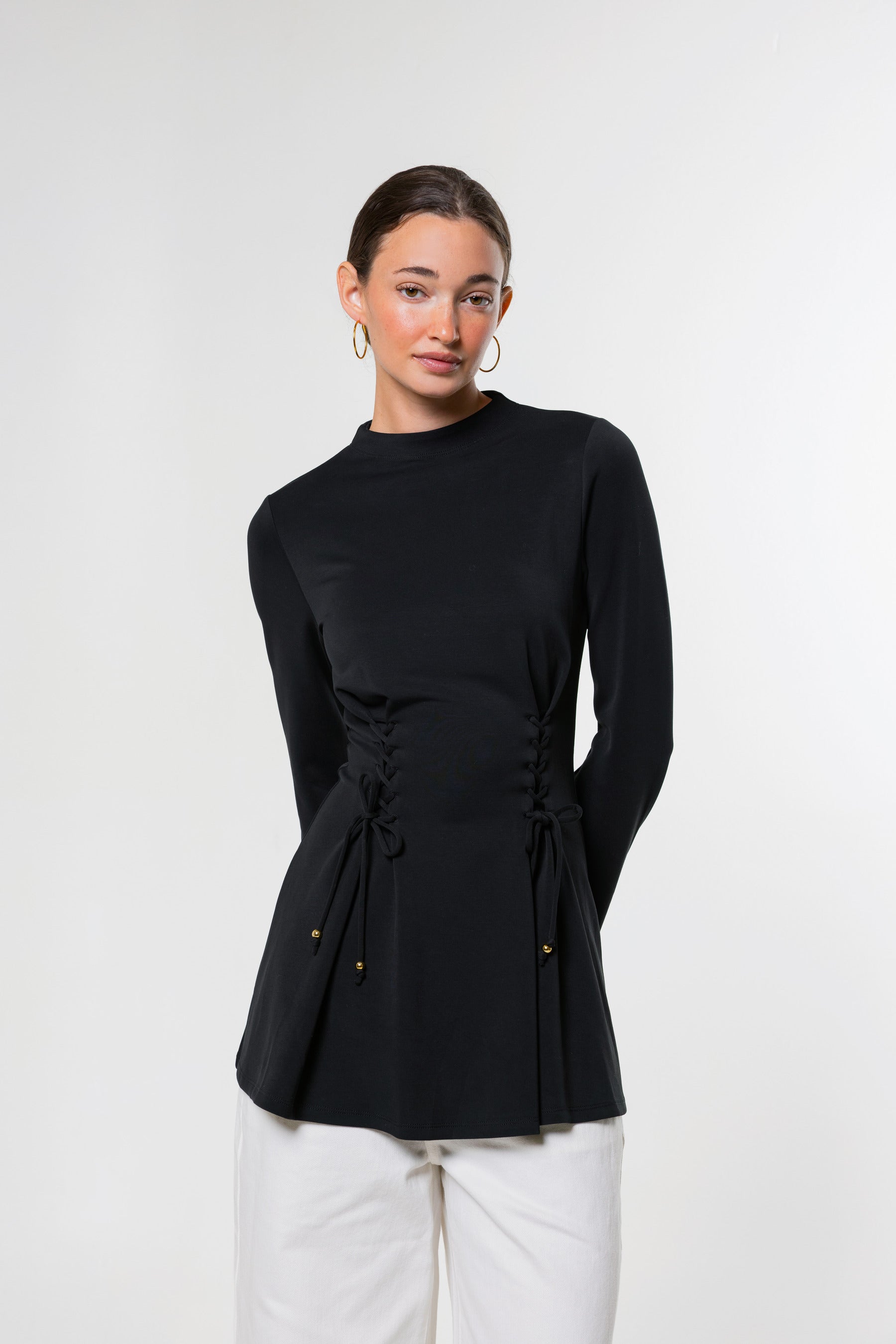 A woman modeling a black long-sleeve mock neck top with a drawstring waist, paired with white pants, against a plain light gray background.