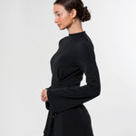 A woman in profile wearing a black long-sleeve wrap-style top with flared cuffs and white pants, photographed against a clean light gray studio background.
