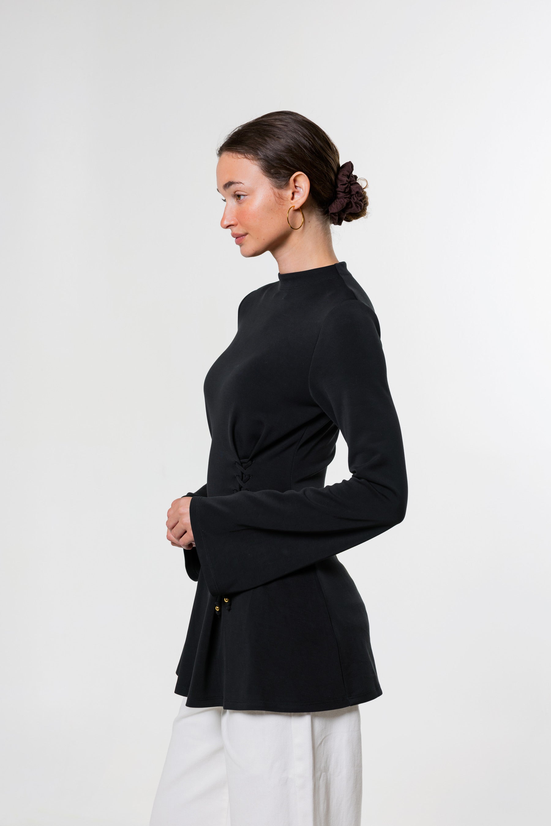 A woman in profile wearing a black long-sleeve wrap-style top with flared cuffs and white pants, photographed against a clean light gray studio background.
