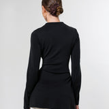 A woman viewed from the back wearing a fitted black long-sleeve top and white pants against a plain light gray studio background.