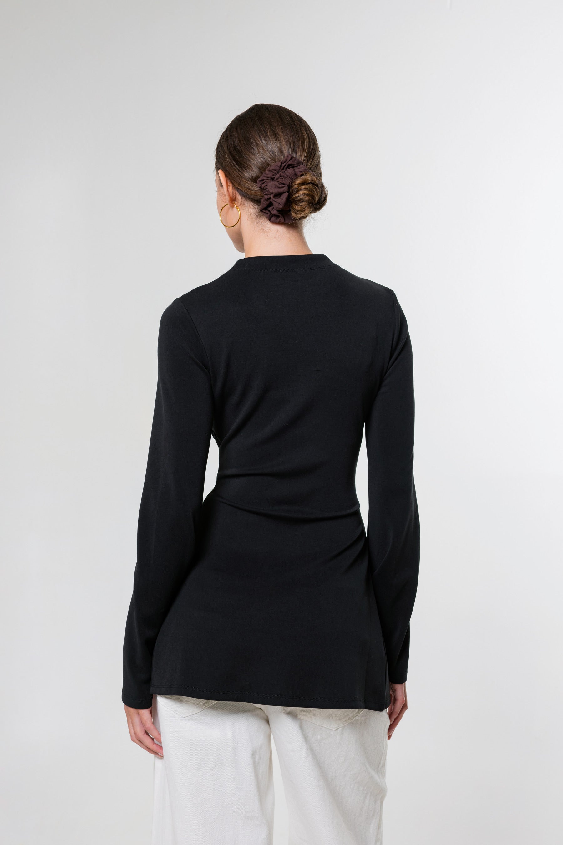 A woman viewed from the back wearing a fitted black long-sleeve top and white pants against a plain light gray studio background.