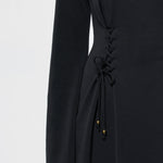 Close-up of a black long-sleeve dress with a lace-up side detail and small gold bead accents, shown on a model against a light gray background.