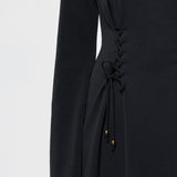 Close-up of a black long-sleeve dress with a lace-up side detail and small gold bead accents, shown on a model against a light gray background.