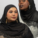 Two women wearing black lace-trimmed hijabs and matching black outer shawls, posed against a light gray studio background.