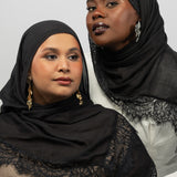 Two women wearing black lace-trimmed hijabs and matching black outer shawls, posed against a light gray studio background.