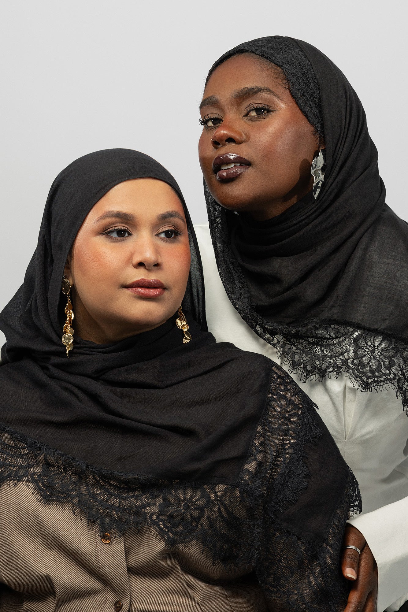 Two women wearing black lace-trimmed hijabs and matching black outer shawls, posed against a light gray studio background.