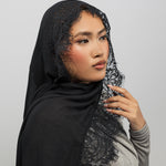 A woman wearing a black lace-trimmed hijab and a gray long-sleeve top, posed against a solid light gray background.