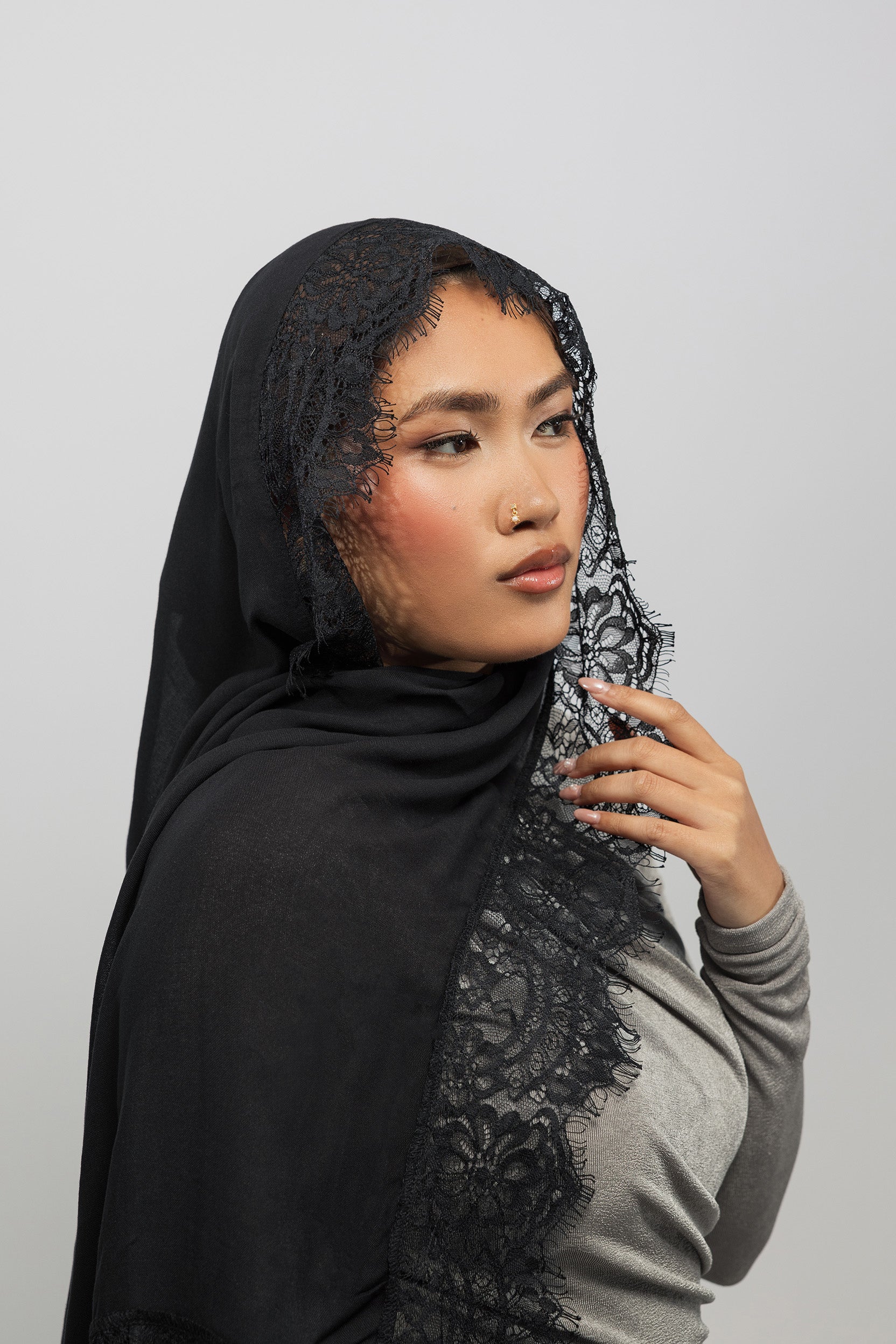 A woman wearing a black lace-trimmed hijab and a gray long-sleeve top, posed against a solid light gray background.