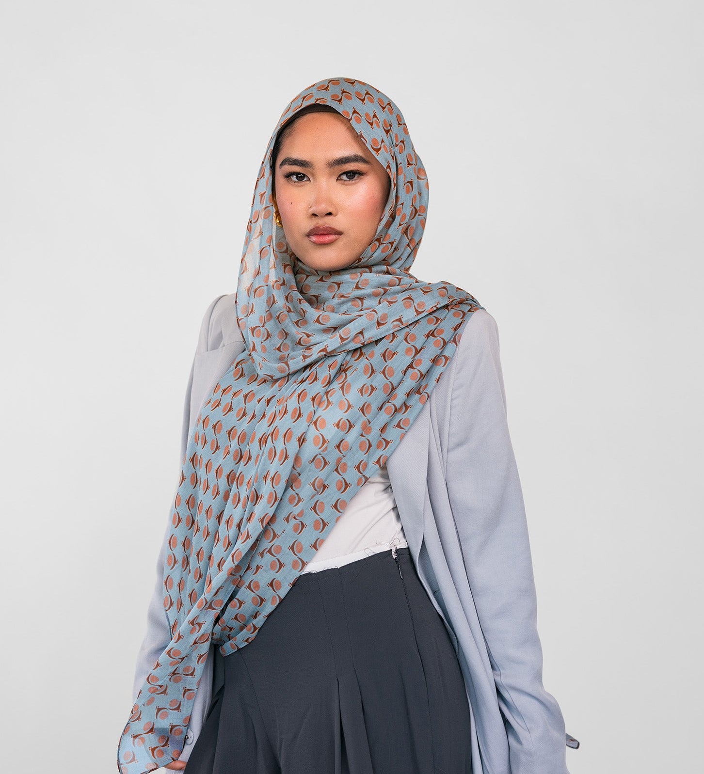 Woman wearing a light blue patterned hijab, light blue cardigan, white top, and dark pants on a white background