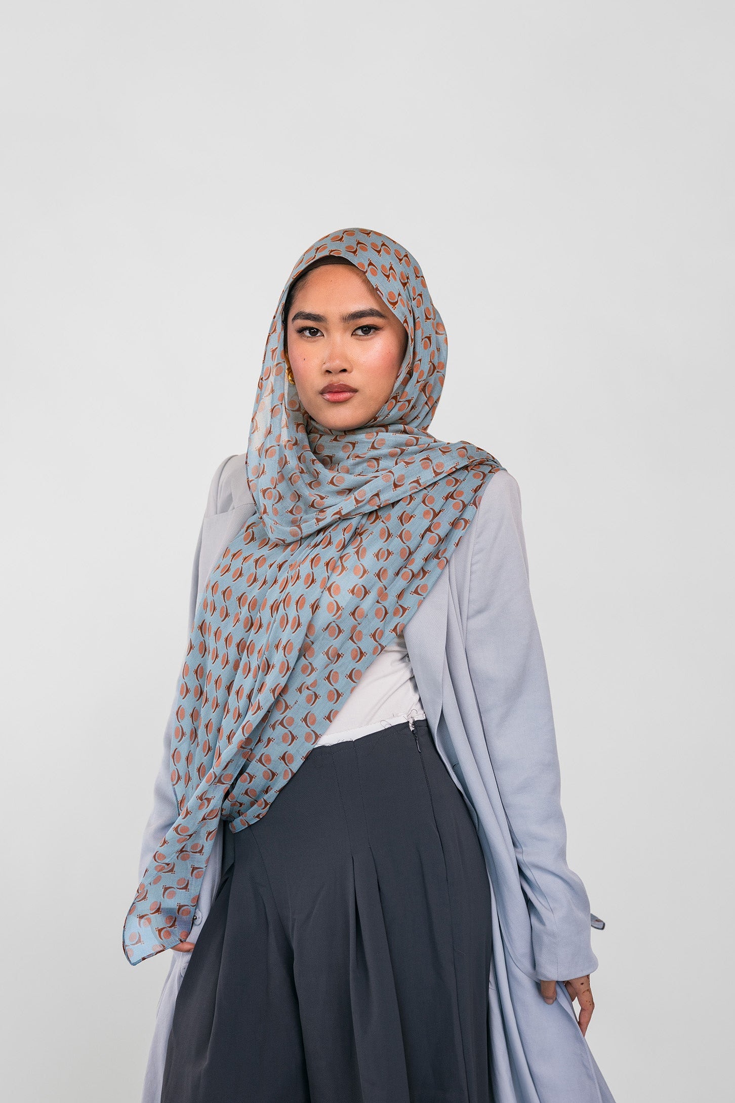 Woman wearing a light blue patterned hijab, light blue cardigan, white top, and dark pants on a white background