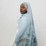 A woman wearing a light blue hijab with intricate white lace trim, posed in profile against a plain light gray studio background.