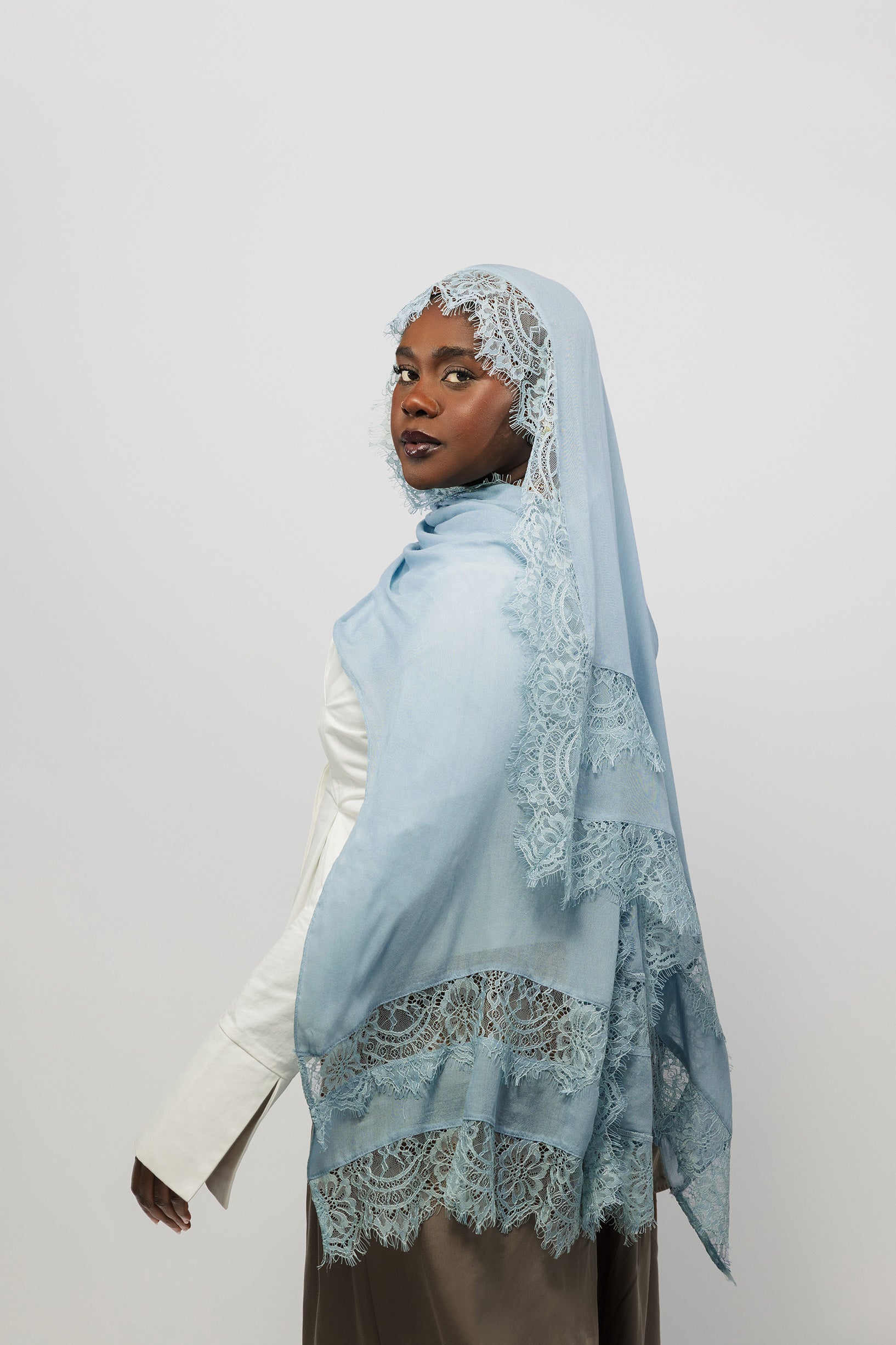A woman wearing a light blue hijab with intricate white lace trim, posed in profile against a plain light gray studio background.