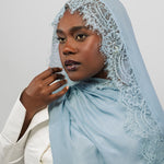 A woman wearing a light blue lace-trimmed hijab and a white blazer, photographed against a plain light gray studio background.