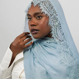 A woman wearing a light blue lace-trimmed hijab and a white blazer, photographed against a plain light gray studio background.