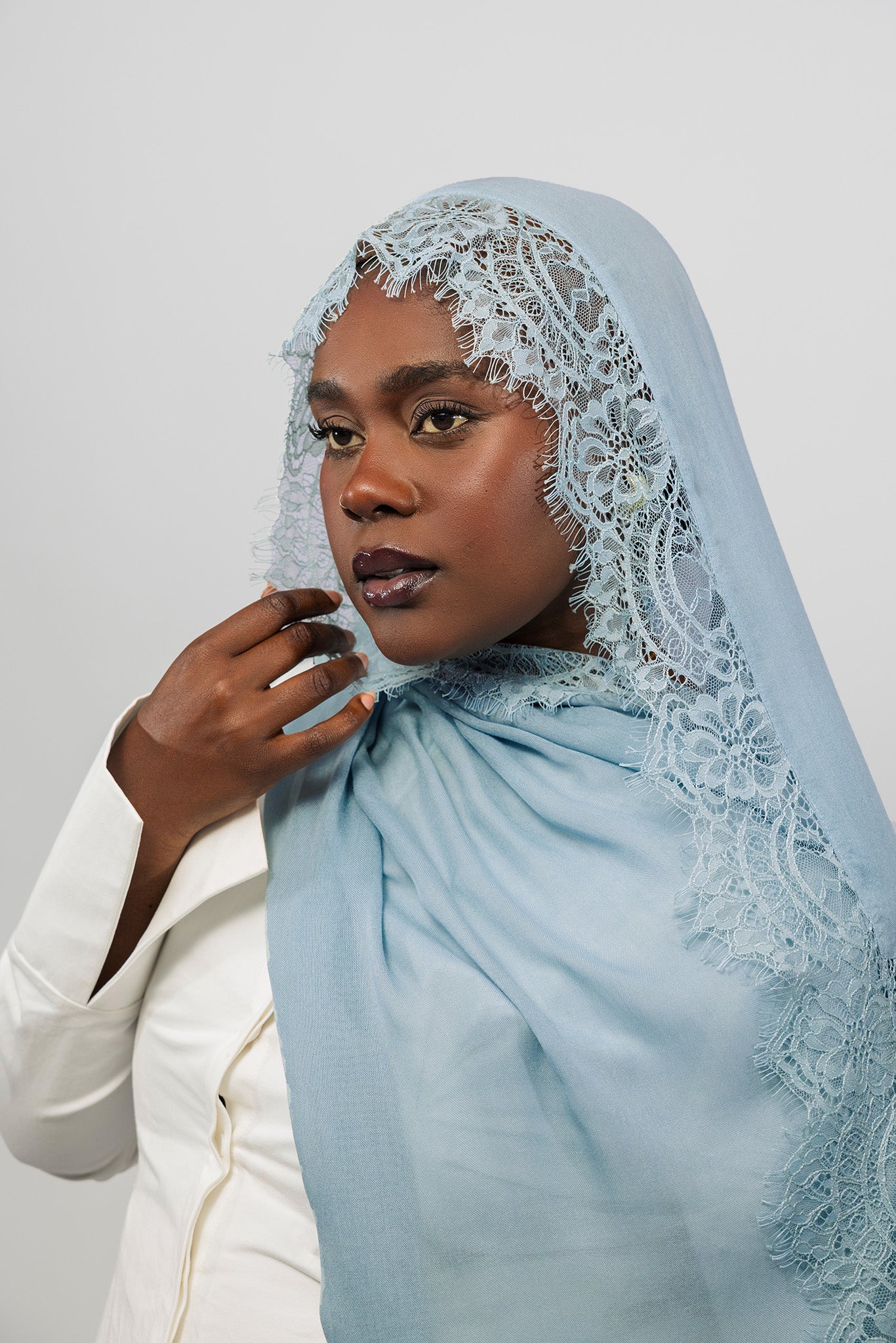 A woman wearing a light blue lace-trimmed hijab and a white blazer, photographed against a plain light gray studio background.