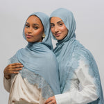 Two women wearing light blue lace-trimmed hijabs and white tops, standing together against a plain light gray studio background.