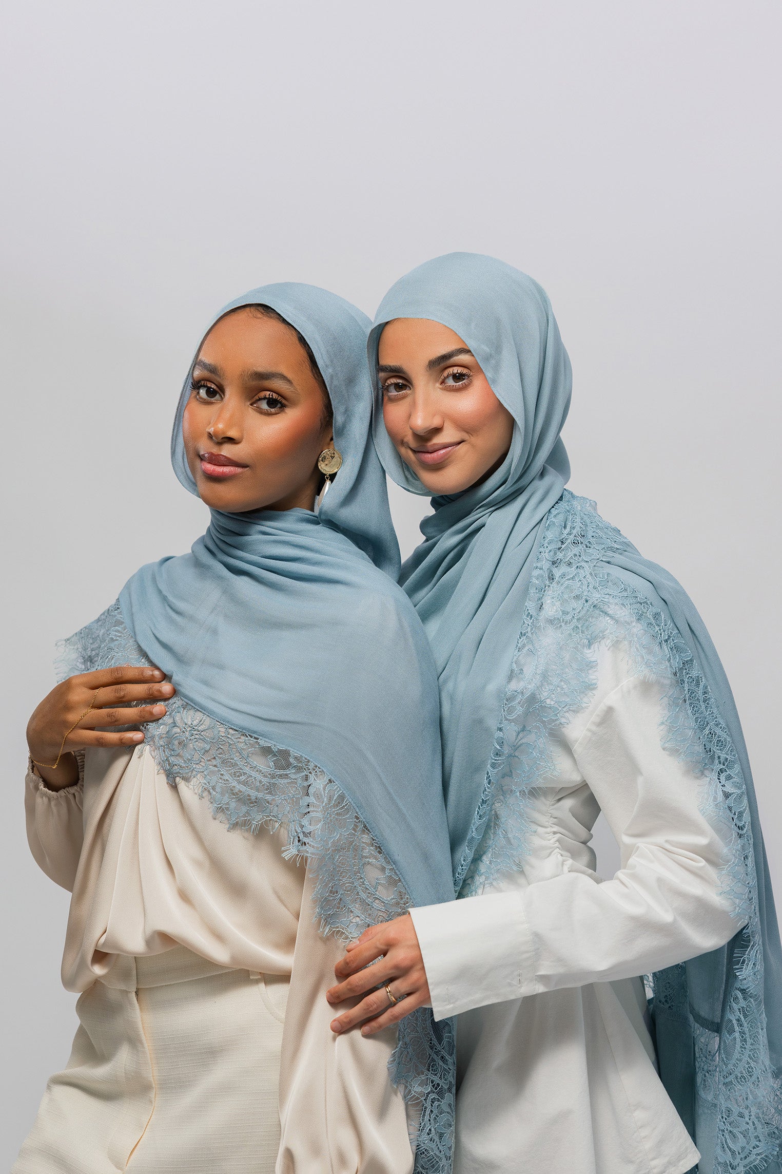Two women wearing light blue lace-trimmed hijabs and white tops, standing together against a plain light gray studio background.