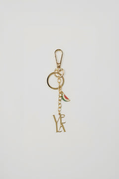Gold keychain with 'VELA' initials and watermelon charm on a light gray background.