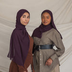 Two women wearing dark purple hijabs and traditional outfits against a plain background