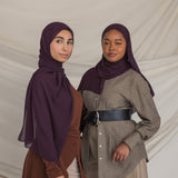 Two women wearing dark purple hijabs and traditional outfits against a plain background