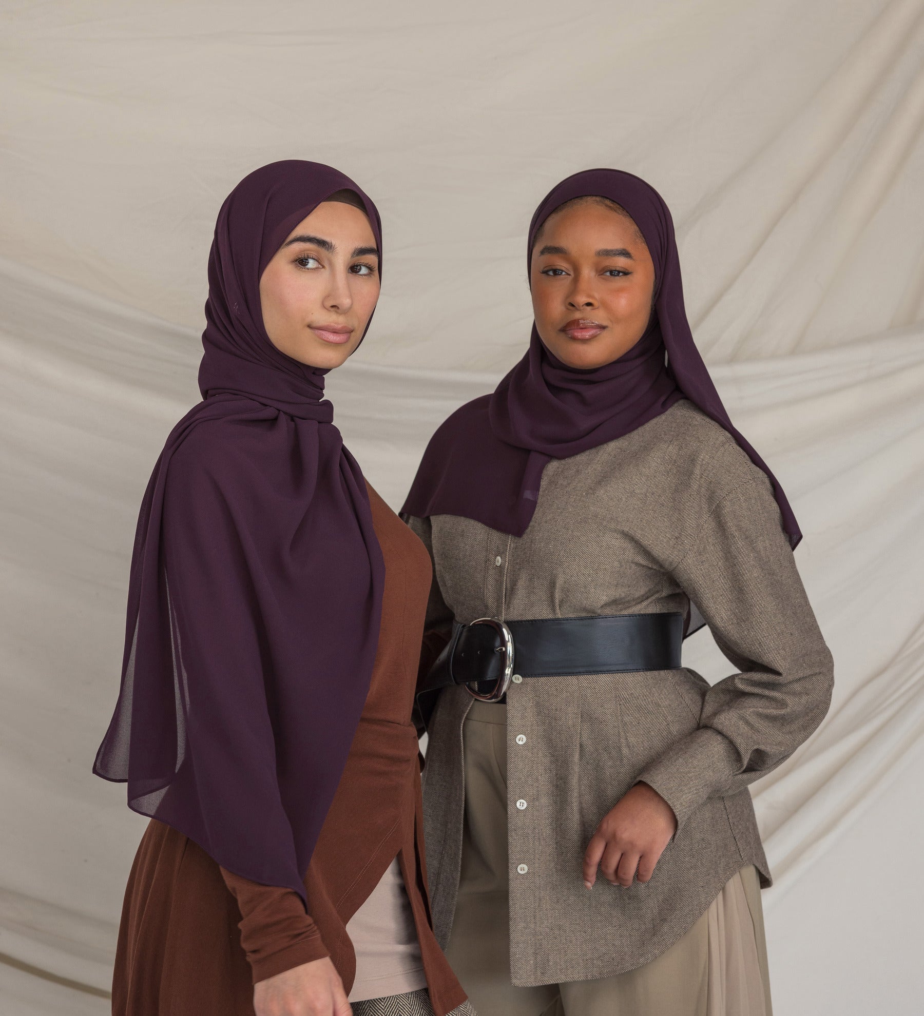 Two women wearing dark purple hijabs and traditional outfits against a plain background