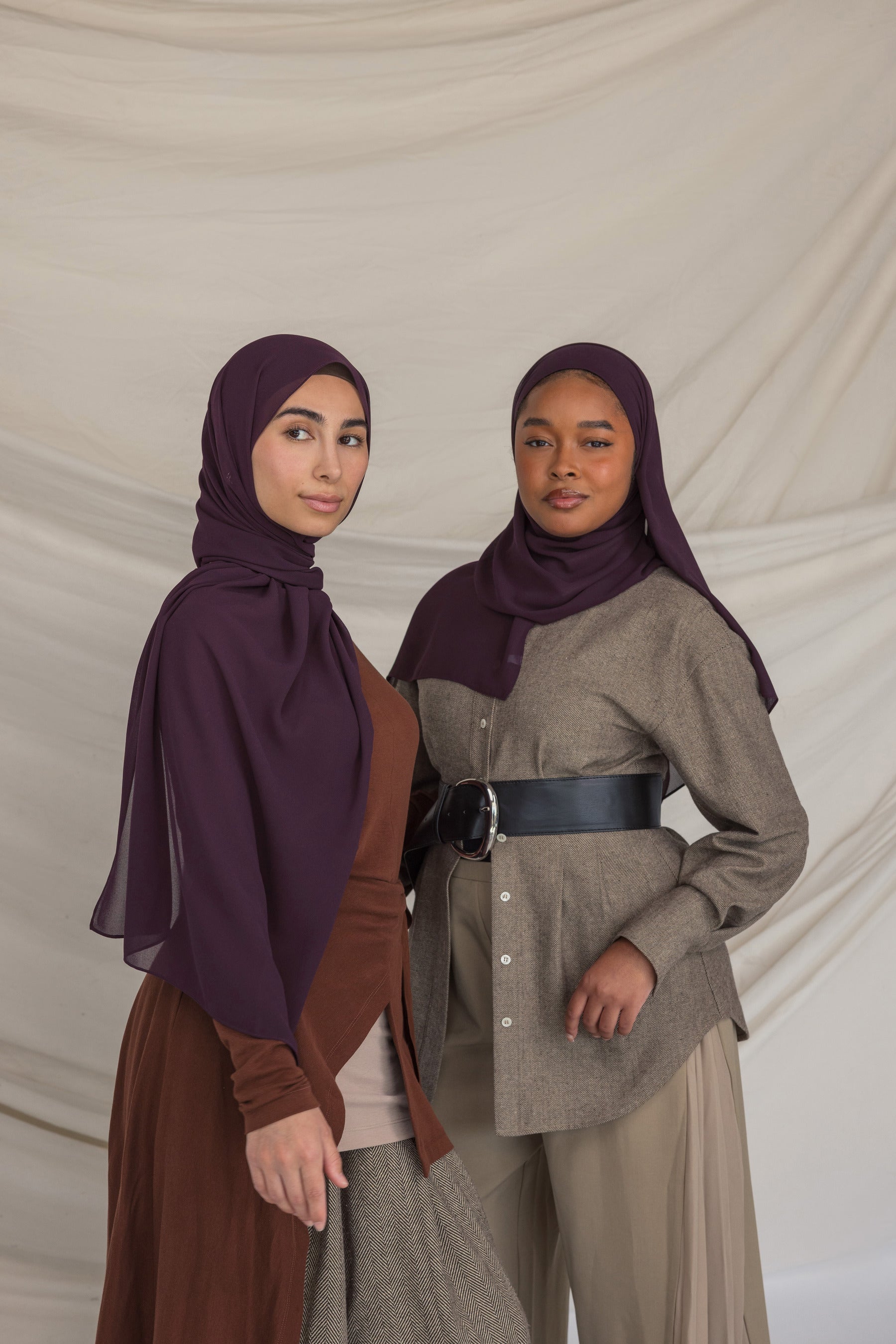 Two women wearing dark purple hijabs and traditional outfits against a plain background