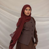 Woman wearing a brown outfit with a red chiffon hijab against a beige background