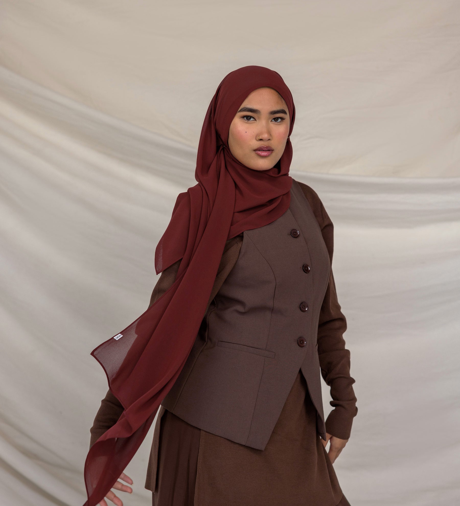 Woman wearing a brown outfit with a red chiffon hijab against a beige background