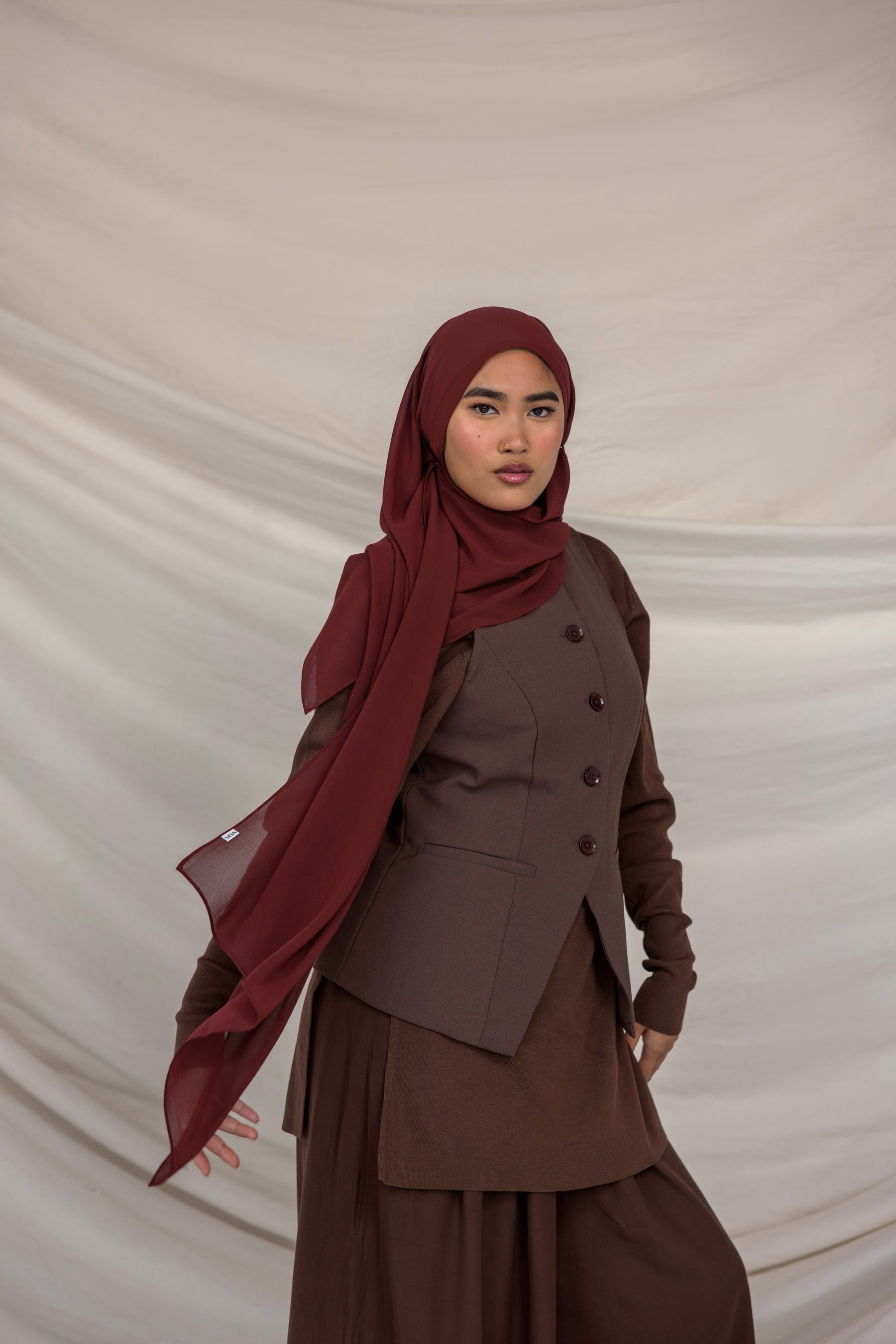 Woman wearing a brown outfit with a red chiffon hijab against a beige background