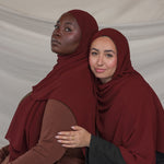 Two women wearing maroon hijabs against a plain background