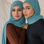 Two women wearing teal chiffon hijabs posing against a neutral background