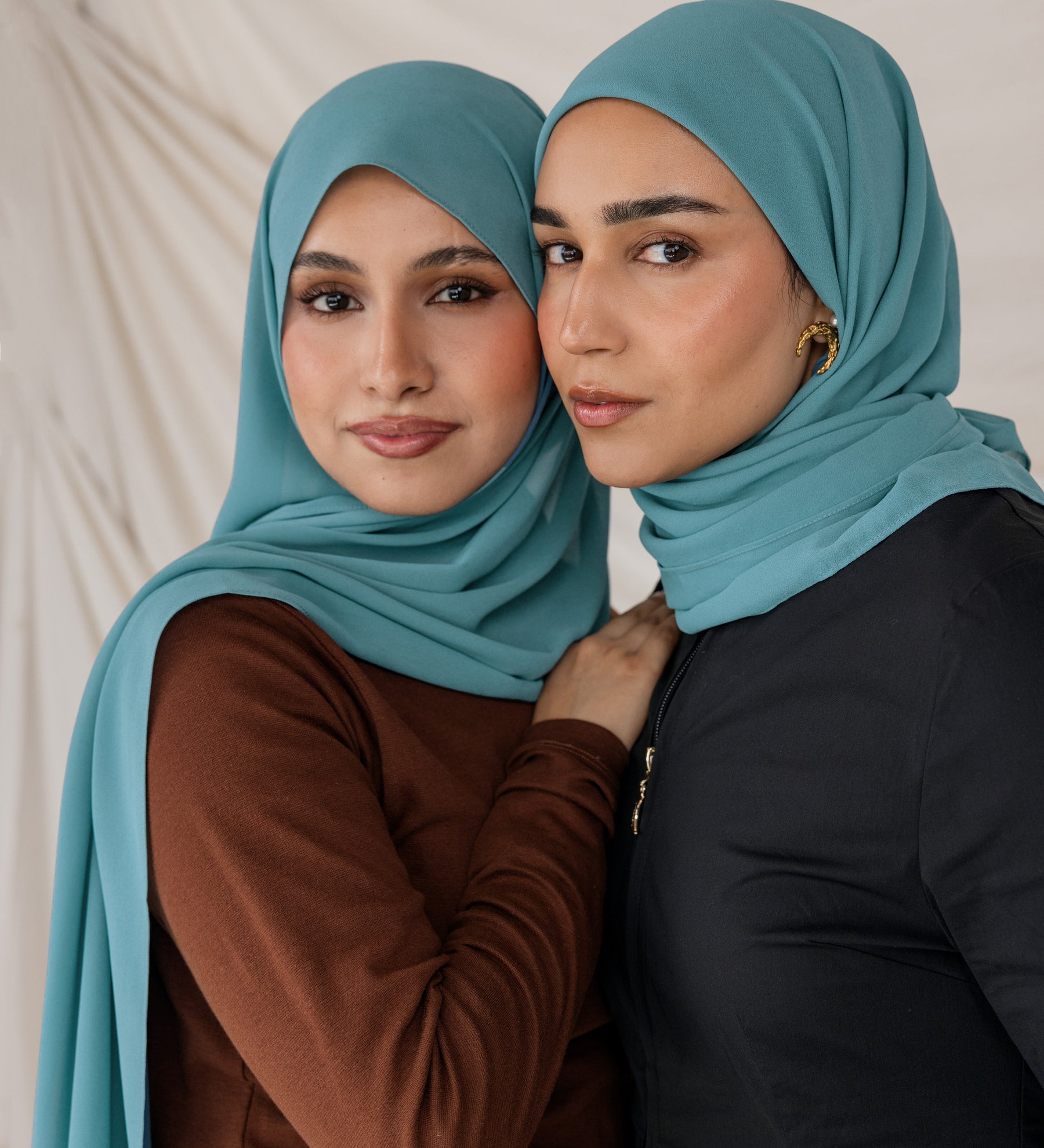 Two women wearing teal chiffon hijabs posing against a neutral background