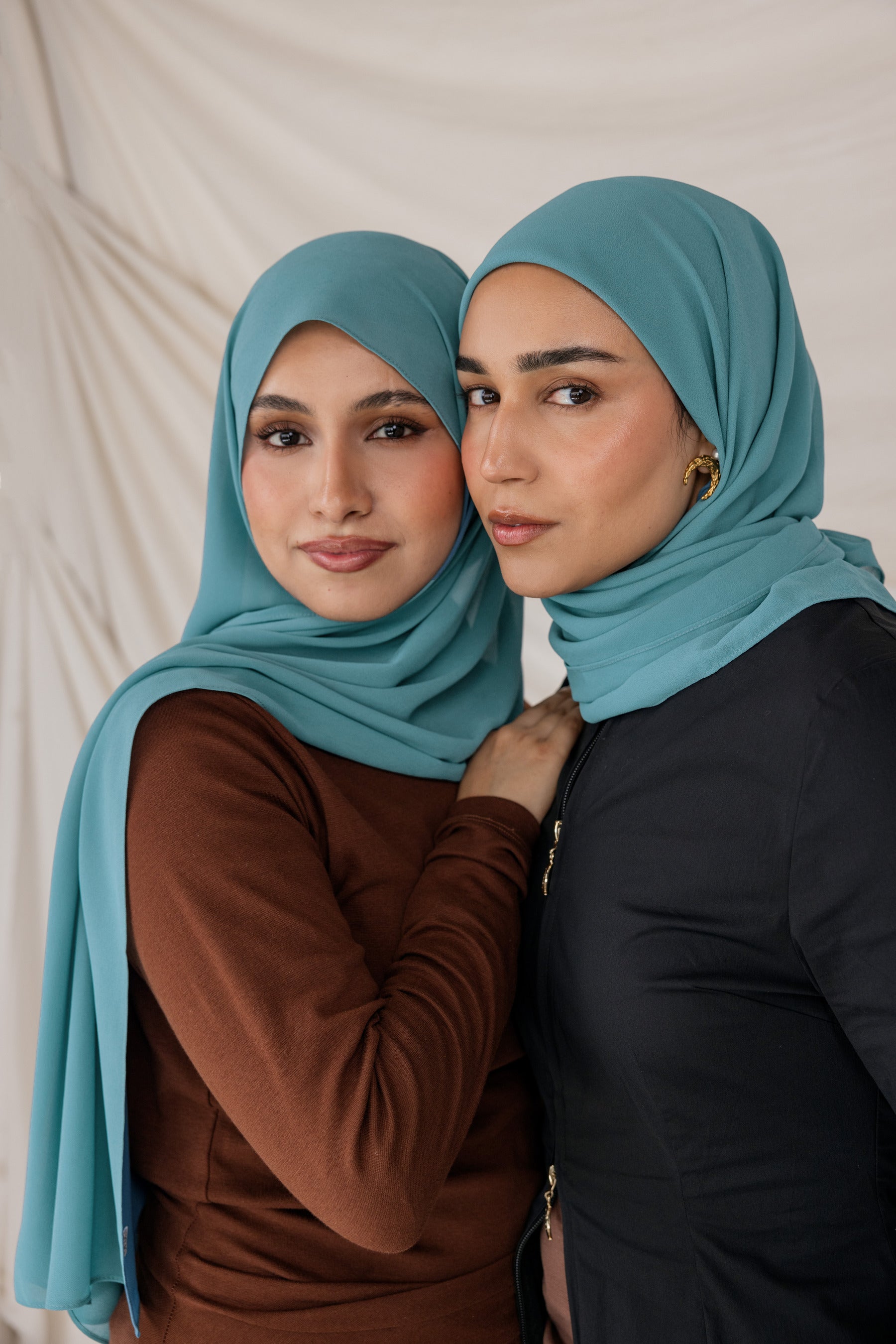 Two women wearing teal chiffon hijabs posing against a neutral background