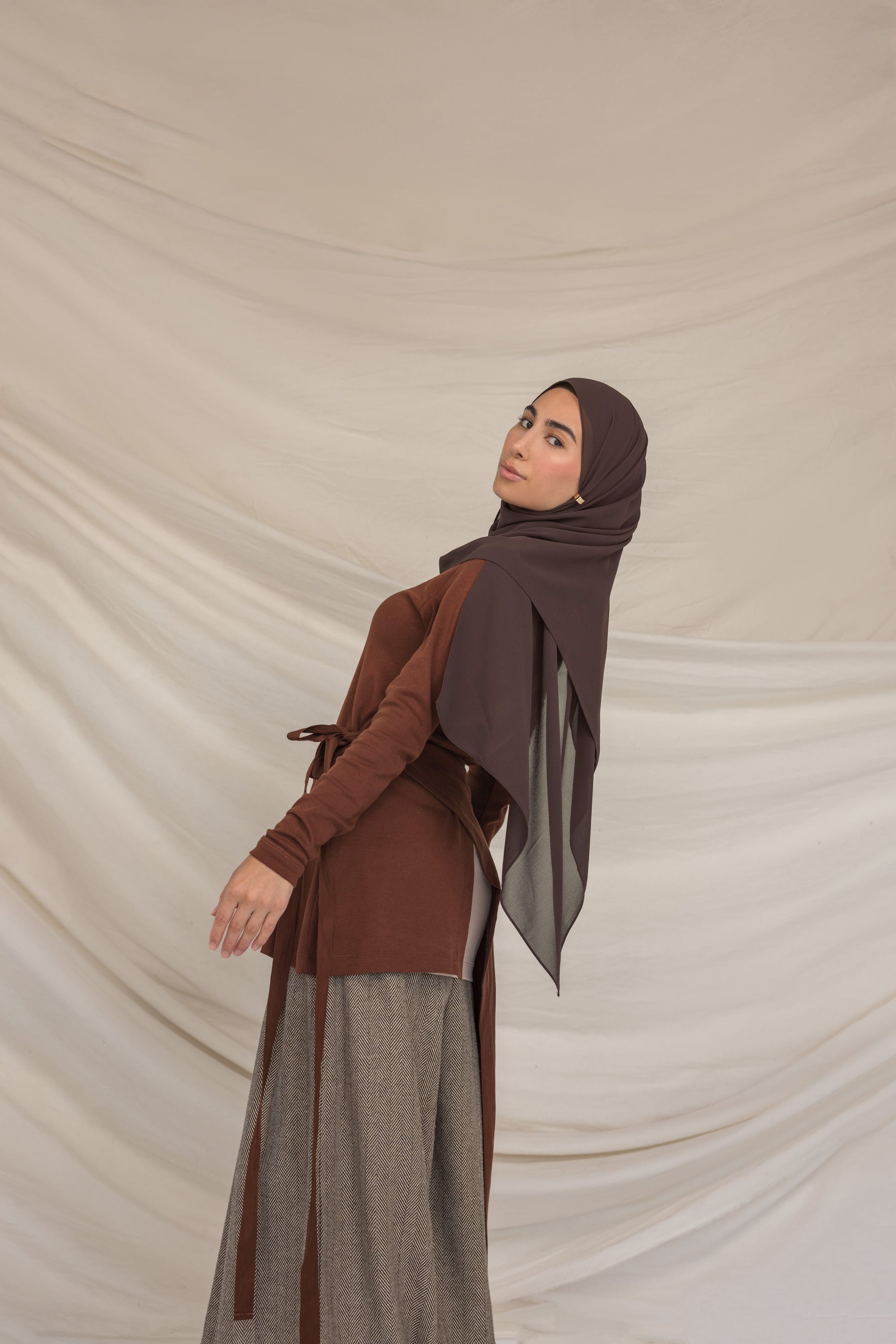 Woman wearing a brown outfit with a brown chiffon hijab against a beige background