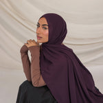 Woman wearing a dark purple hijab and brown long-sleeve top against a beige background